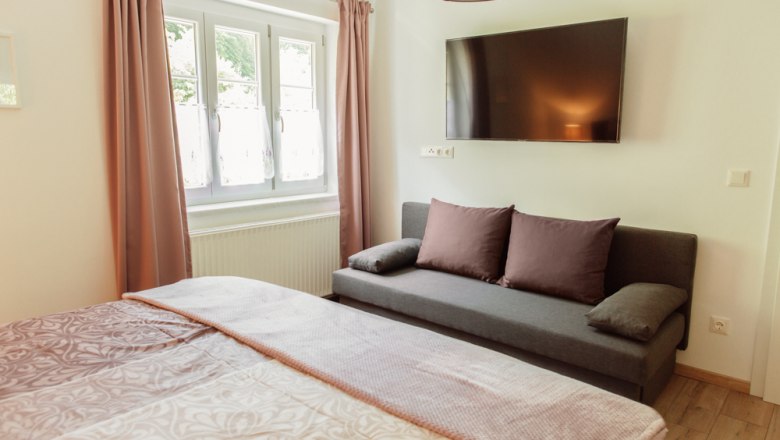 Interior view of a bedroom with bed, sofa, window and wall-mounted TV.