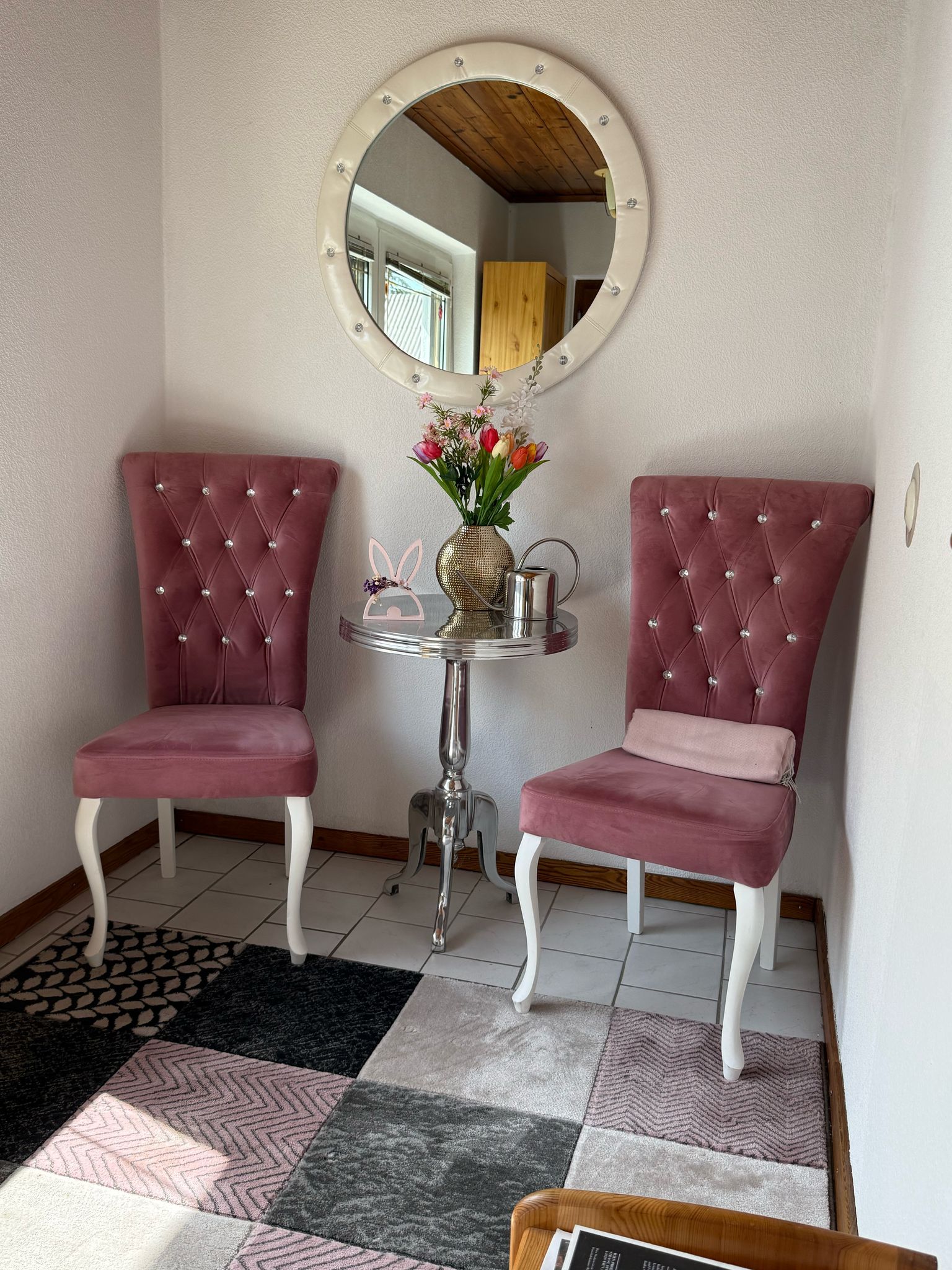 An anteroom with two pink chairs, a round mirror and a small table with flowers.