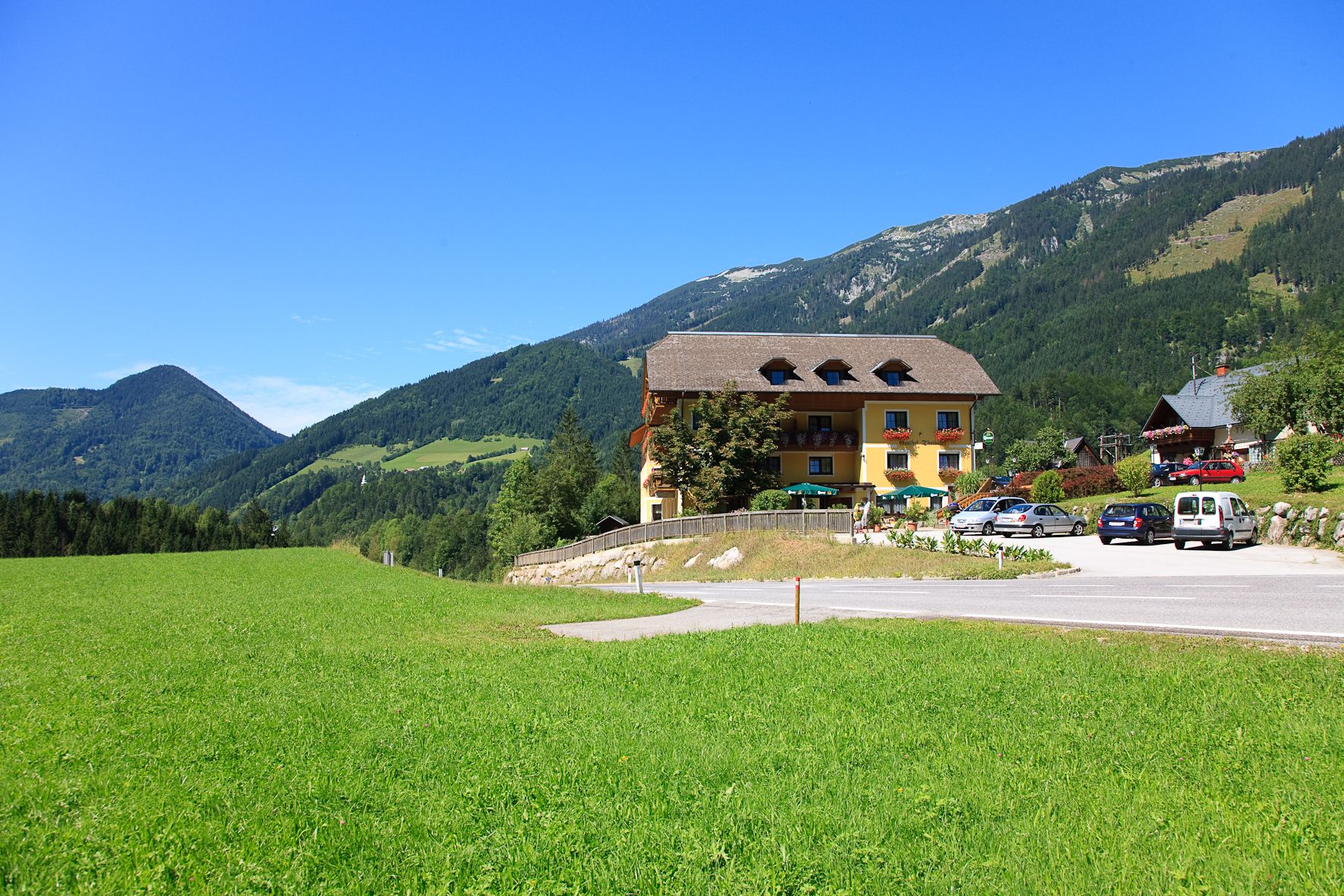 A yellow building with a balcony and flowers, surrounded by green meadows and mountains under a blue sky.