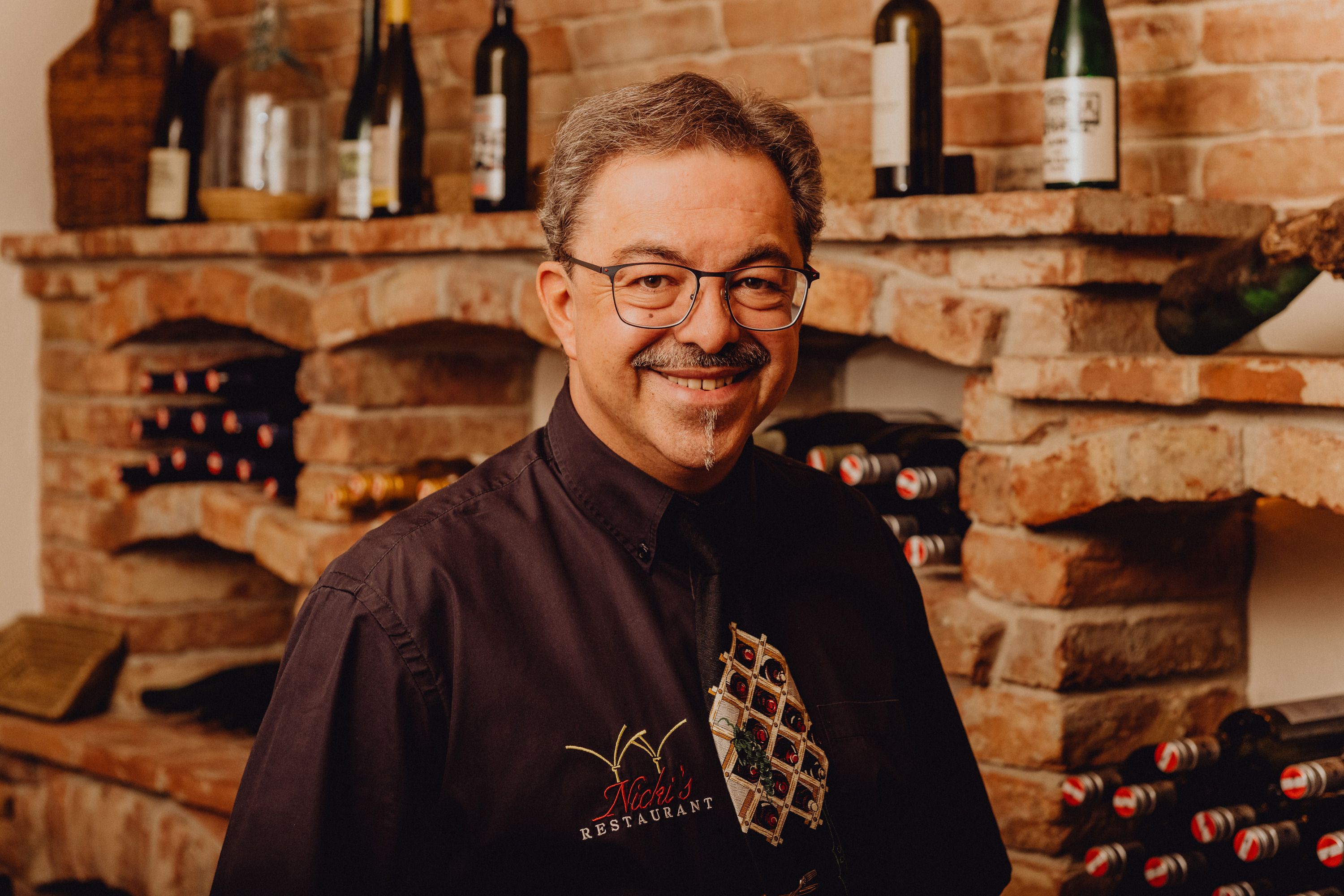 A man with glasses and a moustache stands smiling in front of a wine rack in a restaurant.
