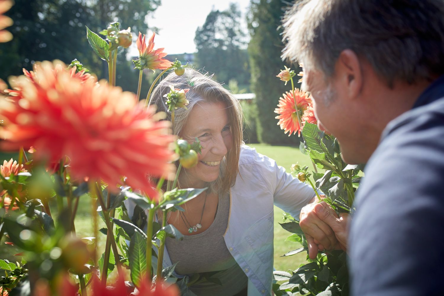 Two people smile at each other between blooming dahlias outdoors.