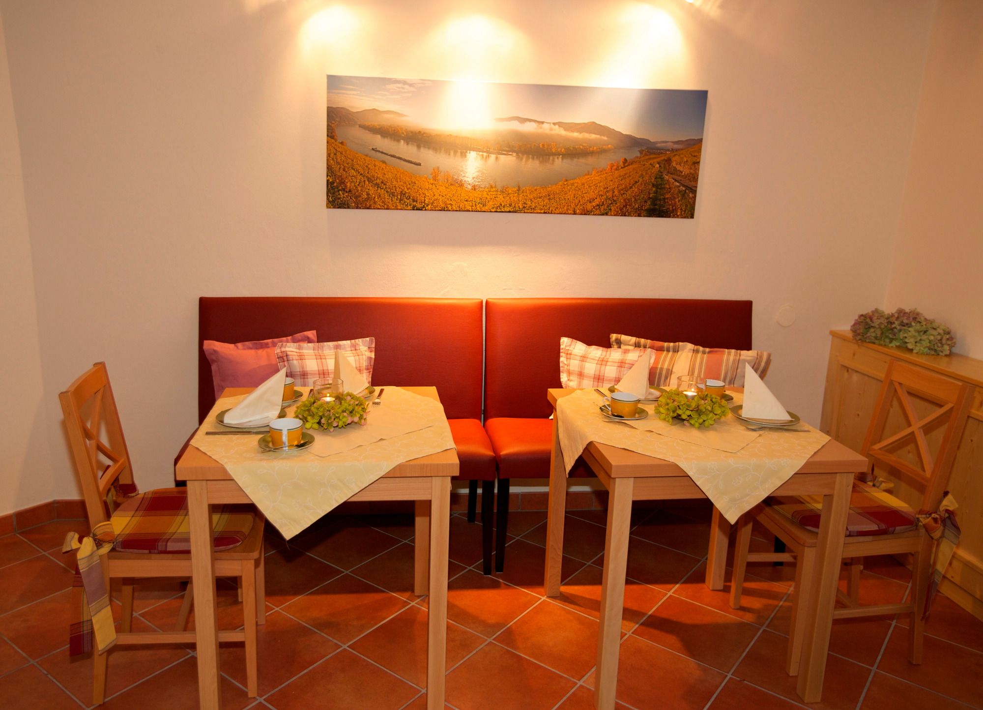 Cozy breakfast room with two set tables, red benches and a landscape painting on the wall.