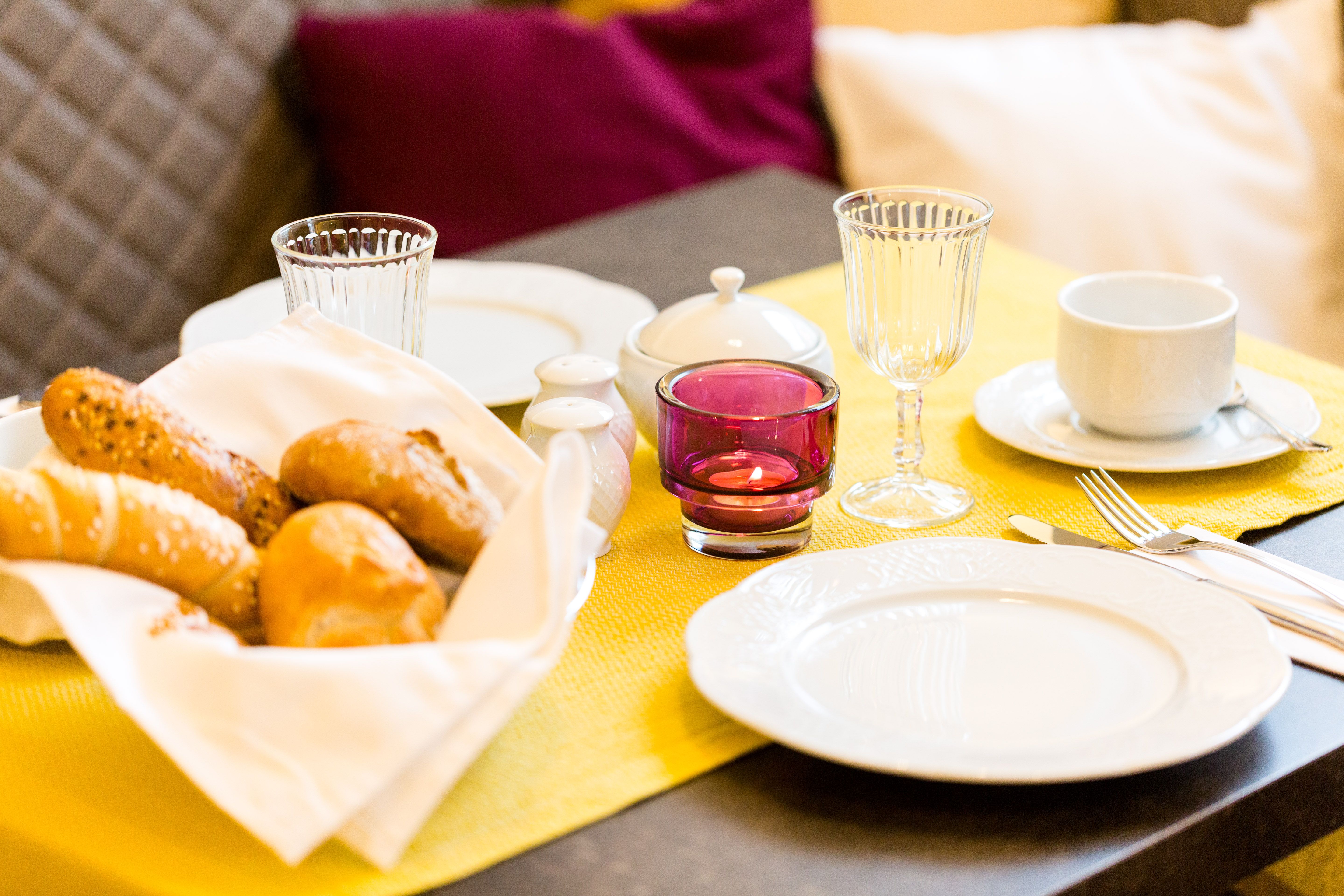 Breakfast table with bread basket, crockery and glasses on a yellow tablecloth.