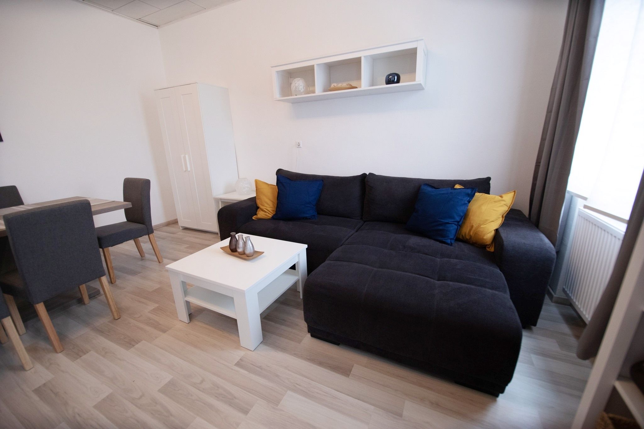 Modern living area with grey sofa, colorful cushions, white coffee table and dining table with chairs.