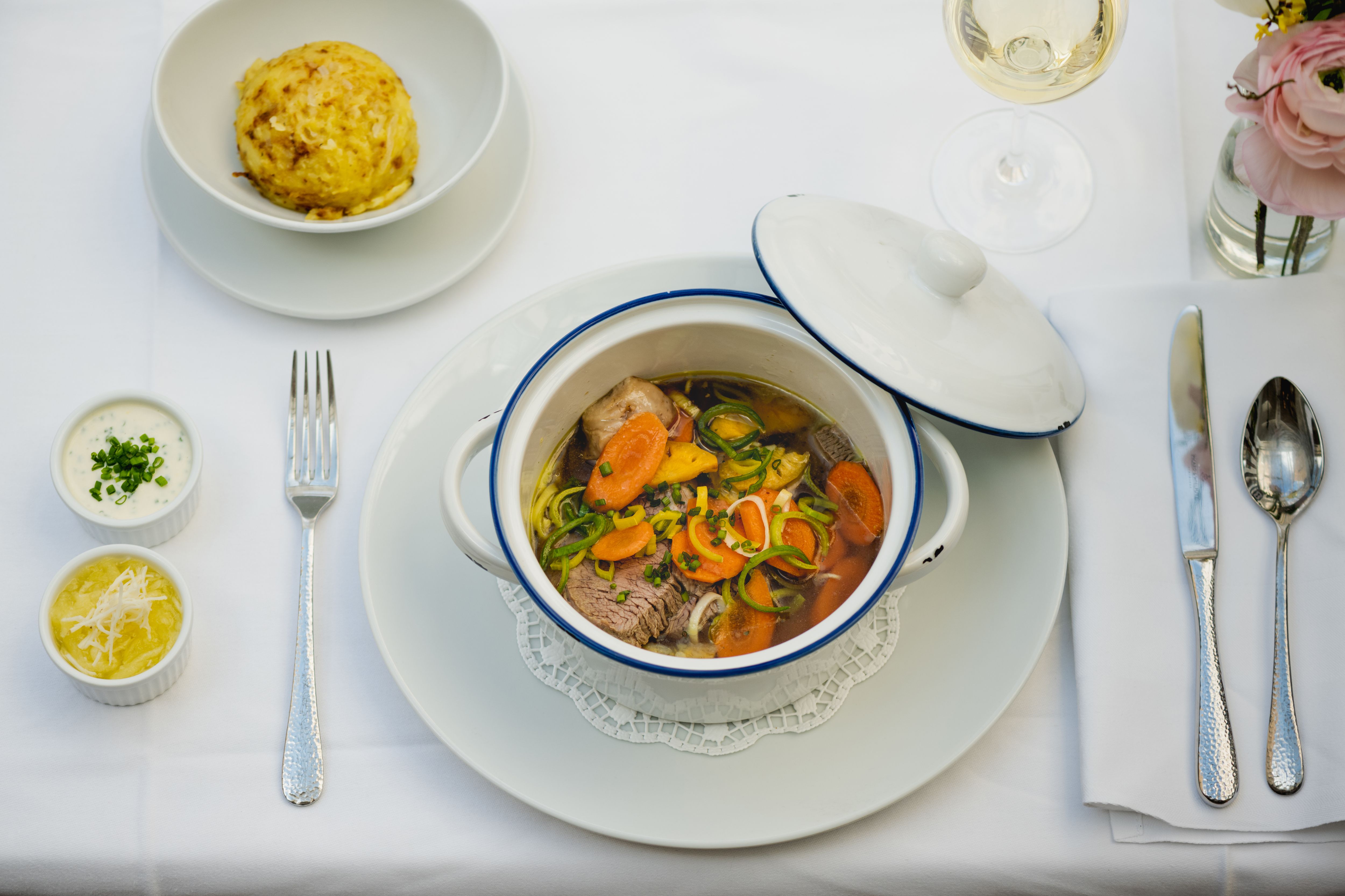 A table set with a stew in a white pot, a dumpling, two sauces and a glass of white wine.