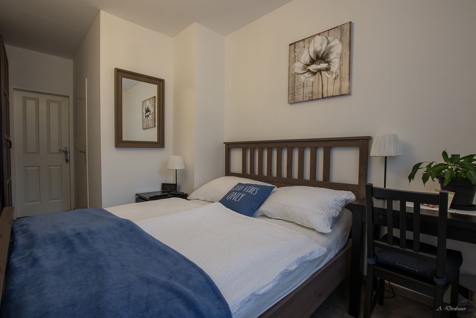 Cozy double room with wooden bed, blue ceiling and decorative cushions. A mirror and a flower picture on the wall, next to it a desk with a plant.