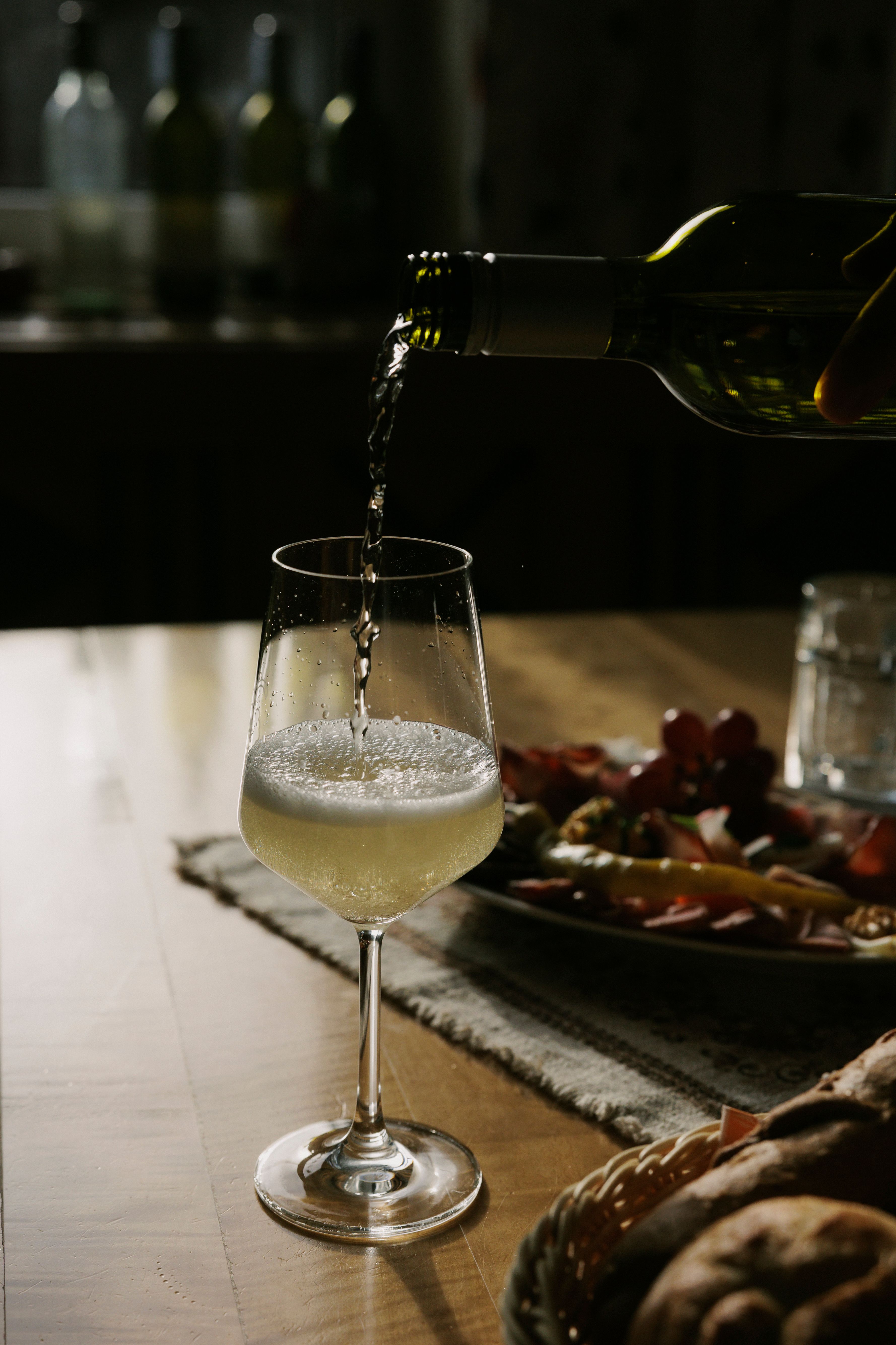 A glass of white wine is poured on a table, with a plate of snacks in the background.