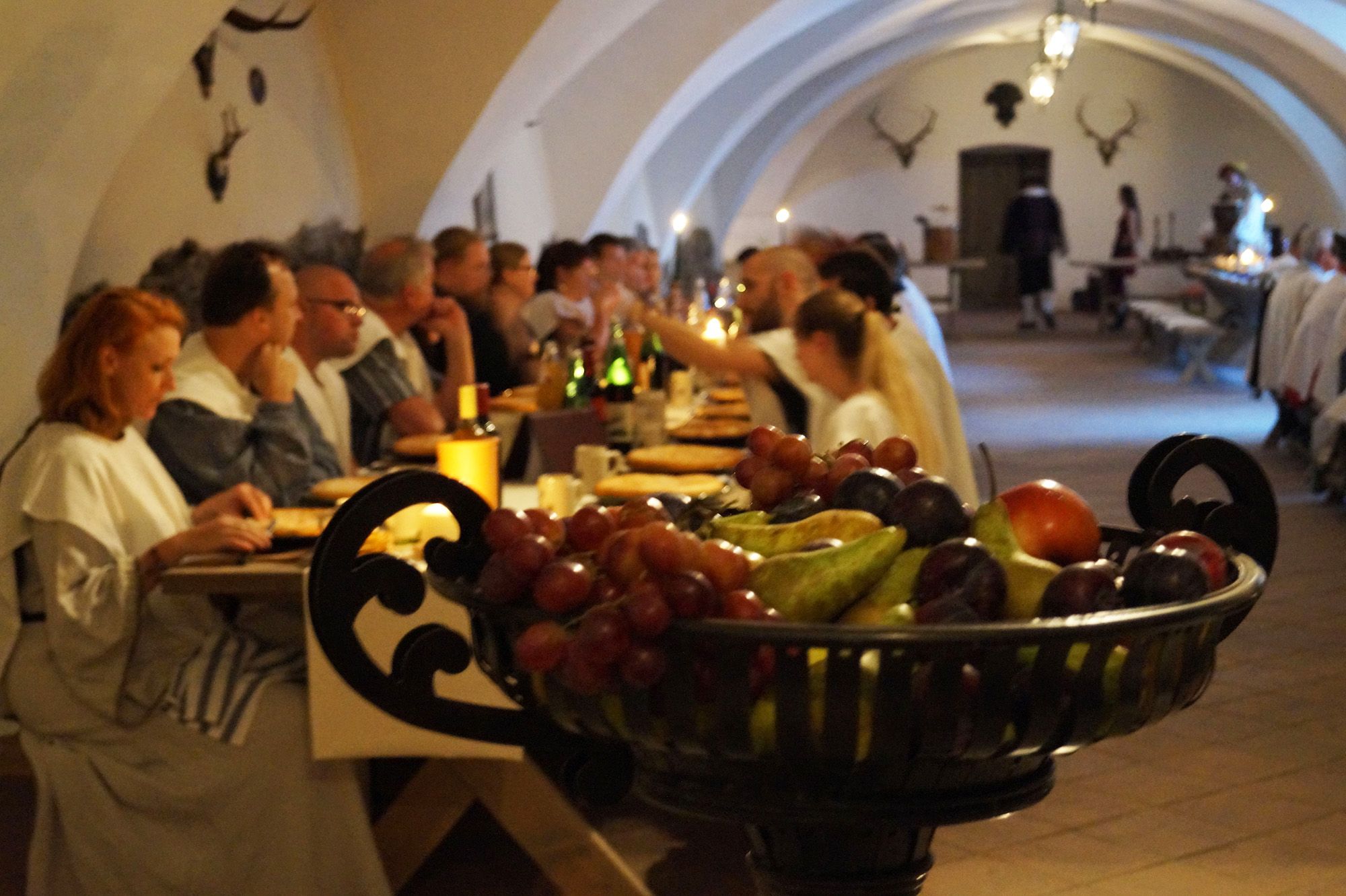 People at a knight's dinner in a vaulted hall with a bowl of fruit in the foreground.