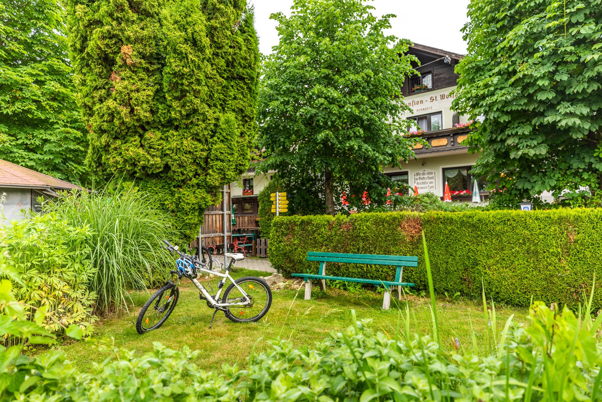 A bicycle is parked next to a bench in the garden of an inn, surrounded by trees and bushes.
