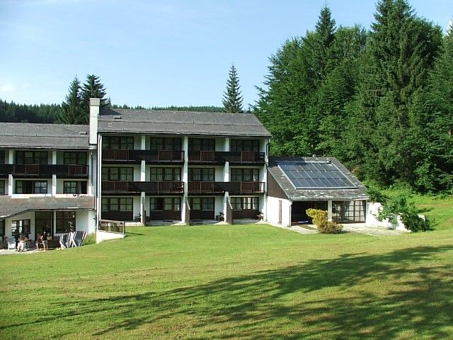 Hotel building with solar panels and green meadow in the foreground, surrounded by forest.