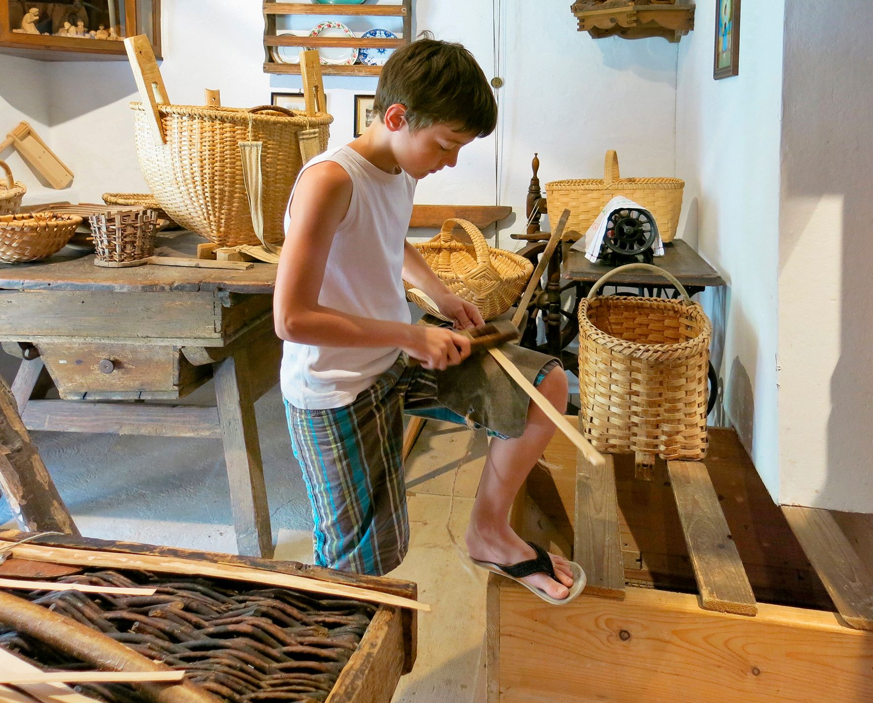 A boy works on a wood project in a museum, surrounded by baskets and traditional tools.