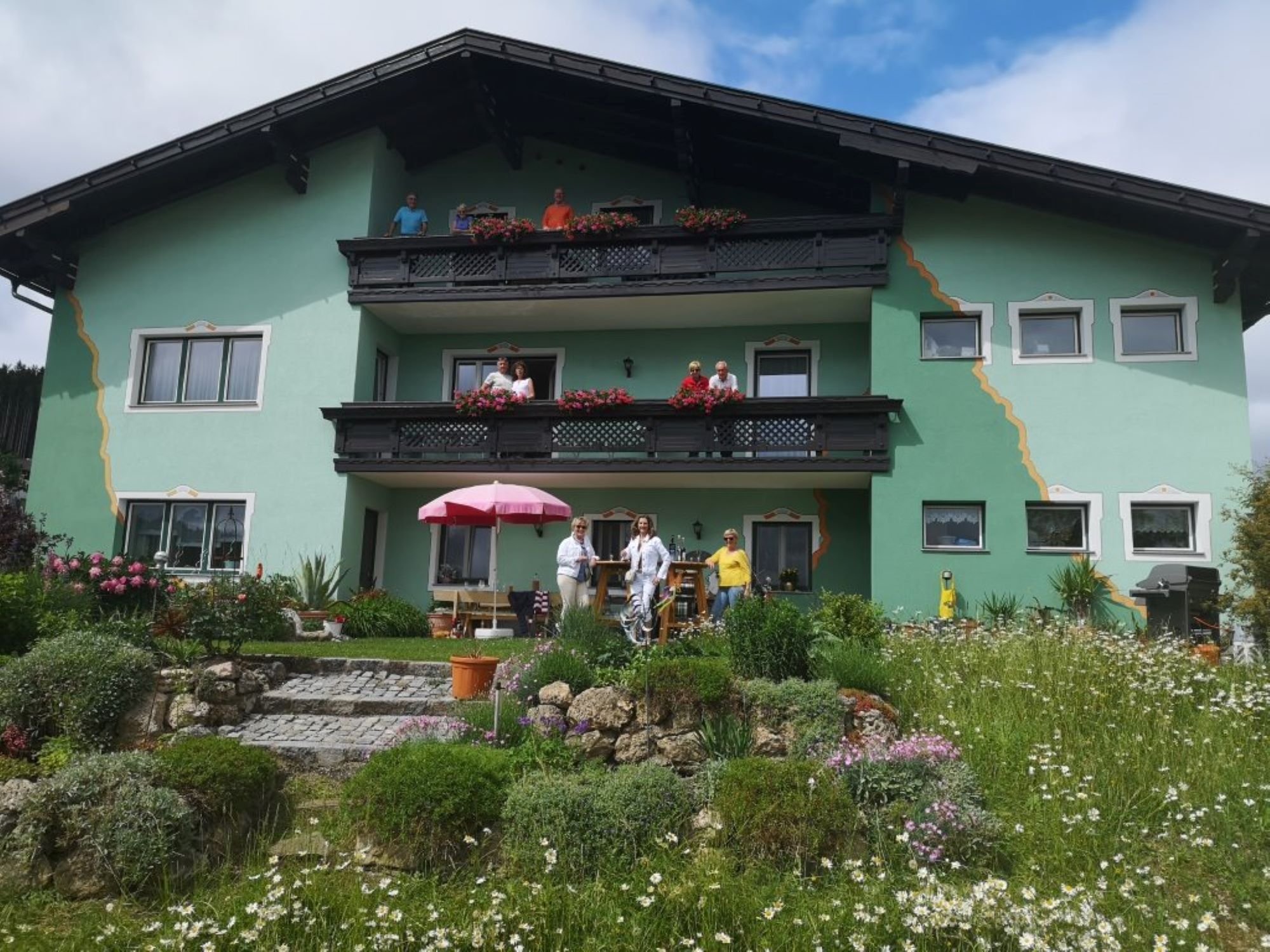 A green house with balconies and people in front of it, surrounded by a flowering garden.