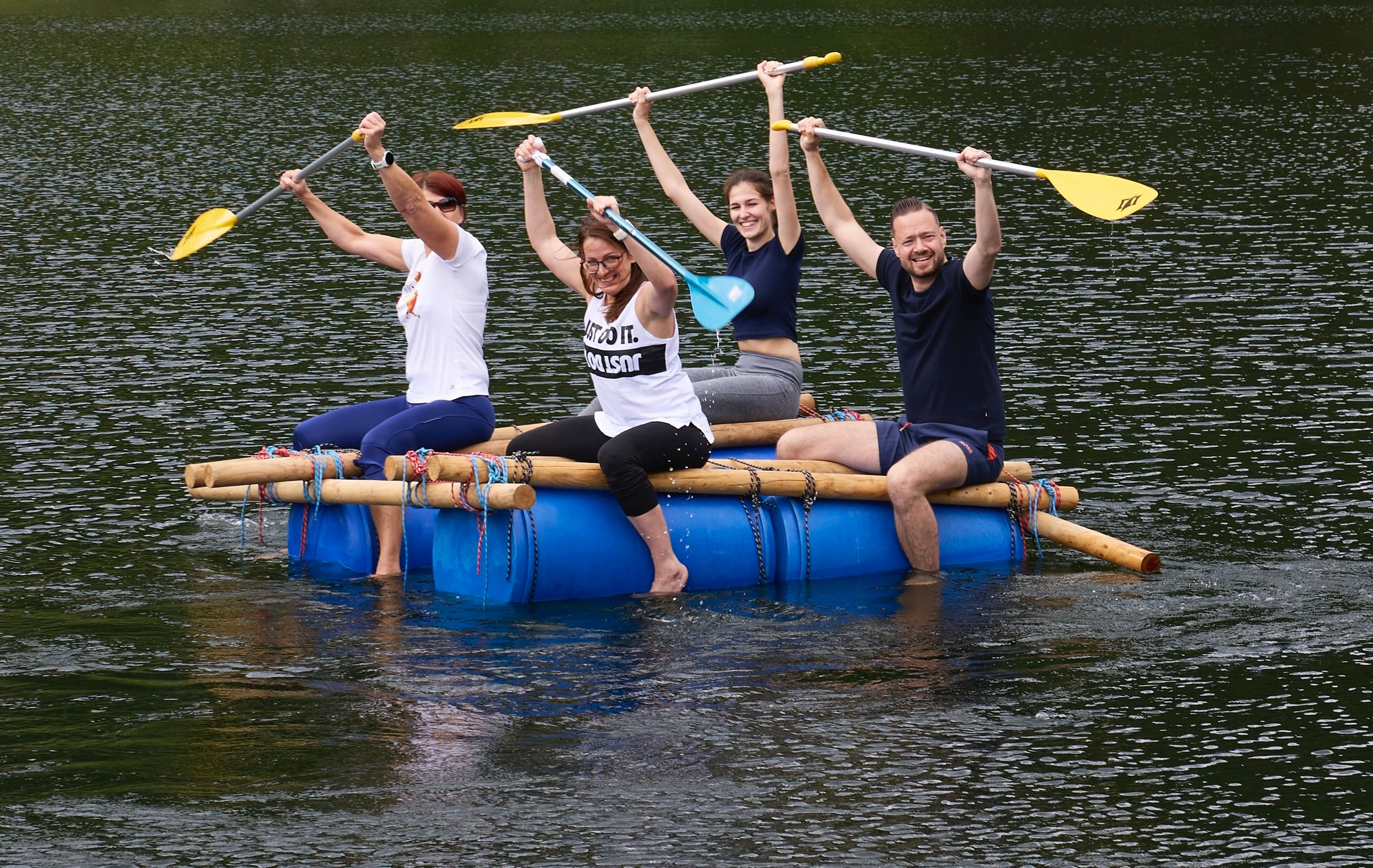 Four people sit on a homemade raft made of blue barrels and wooden poles on a lake and happily lift their paddles into the air.