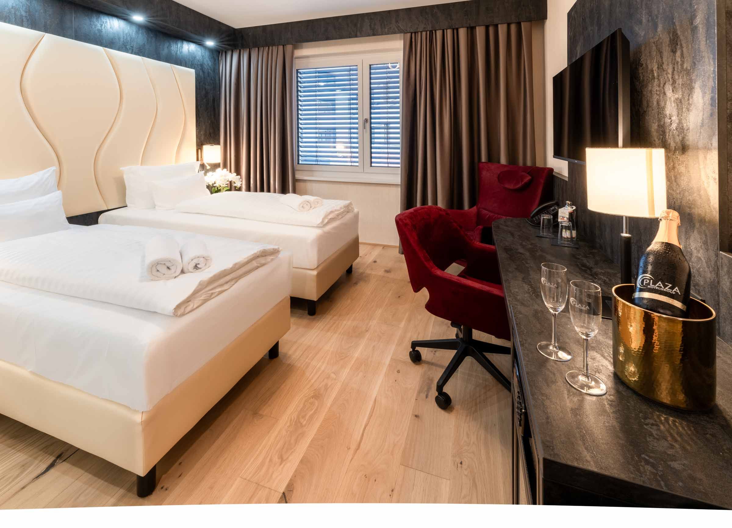 Hotel room with two single beds, red armchair, desk, TV and bottle of champagne.