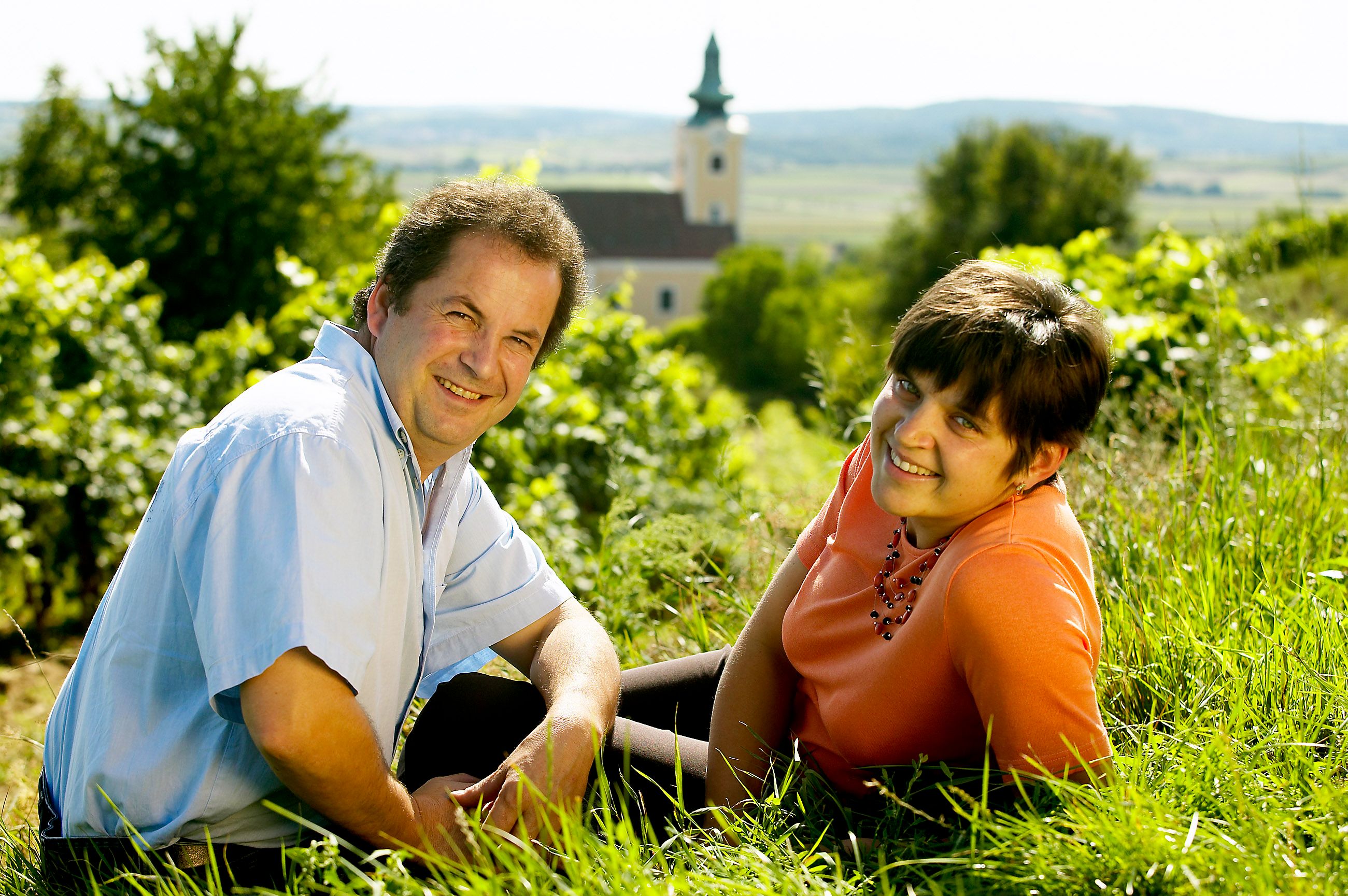 A man and a woman sit smiling on the grass in front of a church in a rural landscape.