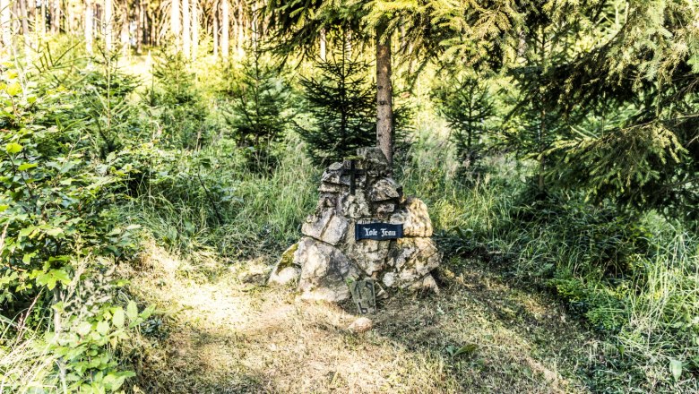 A memorial stone with the inscription 'Dead Woman' stands in a forest, surrounded by trees and grass.