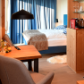 Cozy hotel room with wooden furniture, a bed, table and chairs. There is a coffee machine on a shelf. Blue curtains at the window.