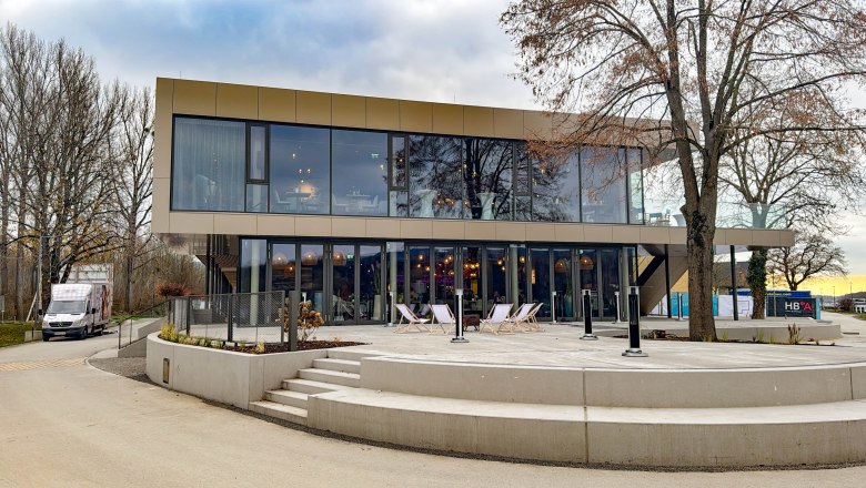 Modern two-storey building with glass façade and terrace, surrounded by trees.