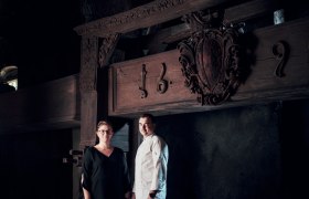 Two people stand in front of an old wooden beam with carvings and the date 1692.