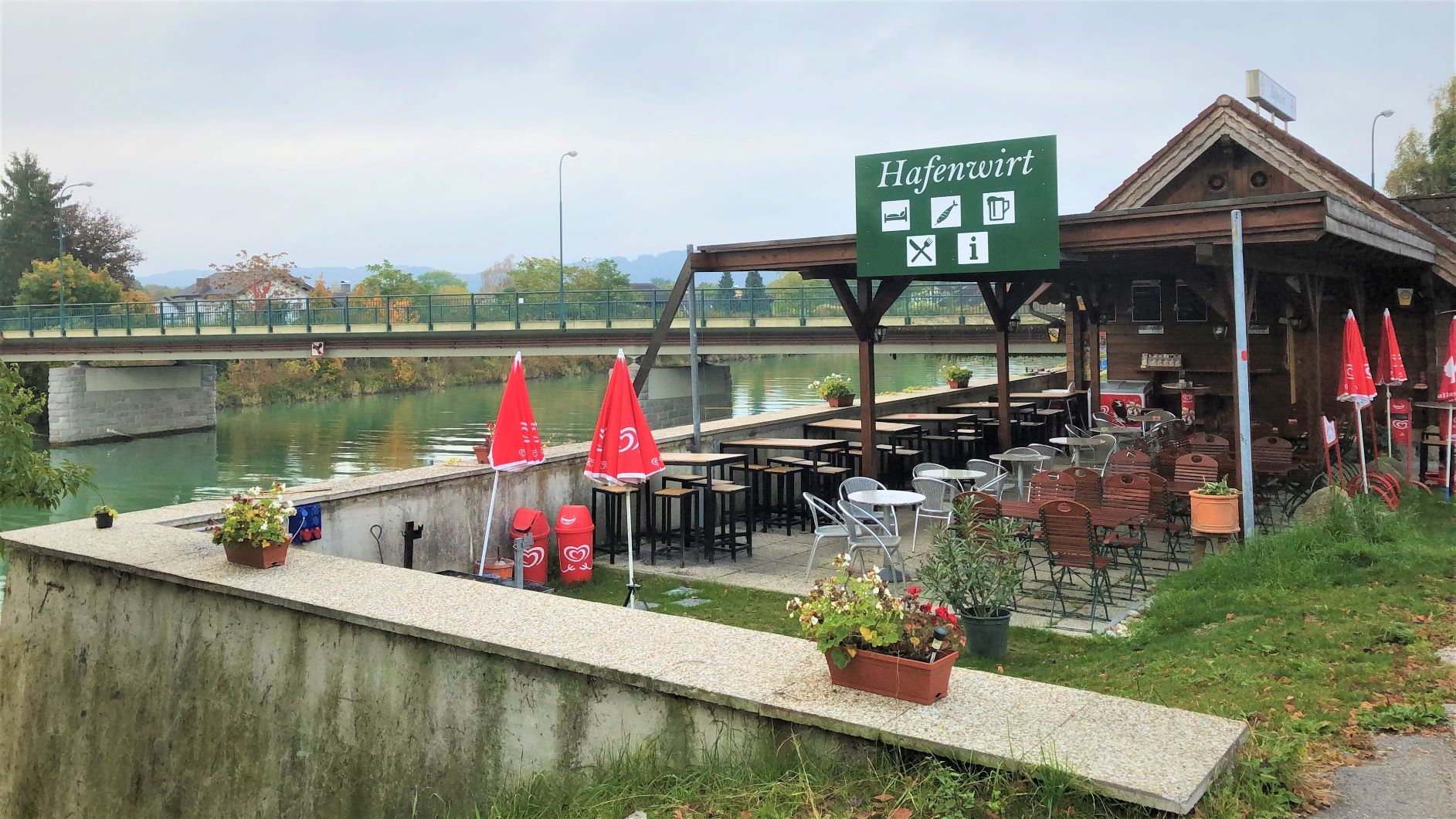A restaurant called Hafenwirt with a terrace and red parasols on a riverbank near a bridge.