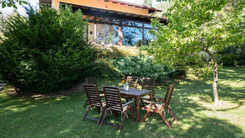 A garden with a wooden table and chairs in front of a vacation home with large windows and surrounded by trees and shrubs.
