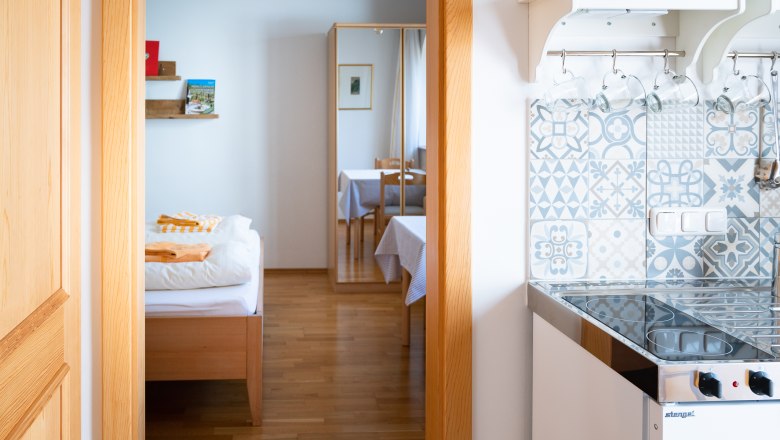 Interior view of a room with bed, mirror, table and small kitchen with patterned tiles.