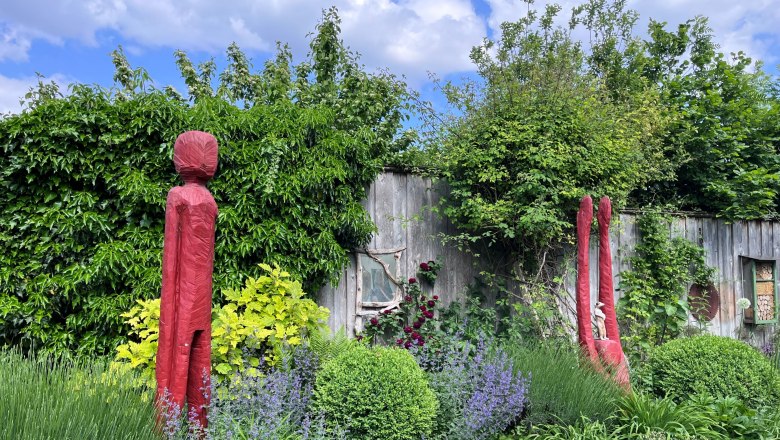 Garden with red sculptures, surrounded by green plants and a wooden wall in the background.