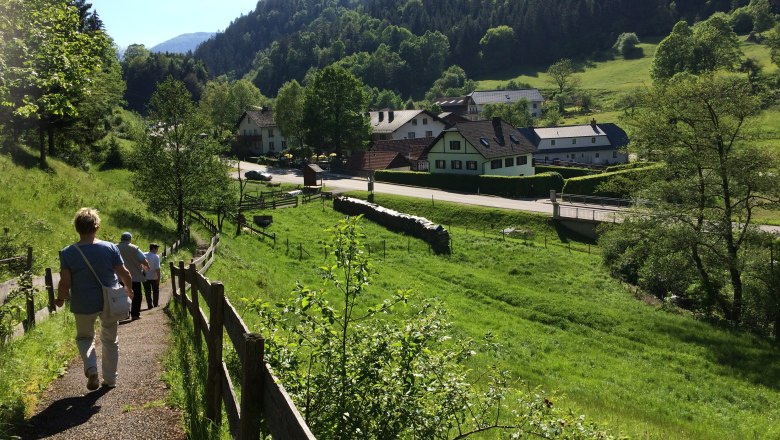People walk along a path in a green, hilly landscape with houses in the background.