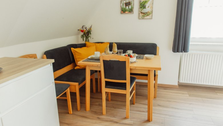 Dining area with wooden table, chairs and corner bench, set with crockery, bouquet of flowers and decoration on the wall.