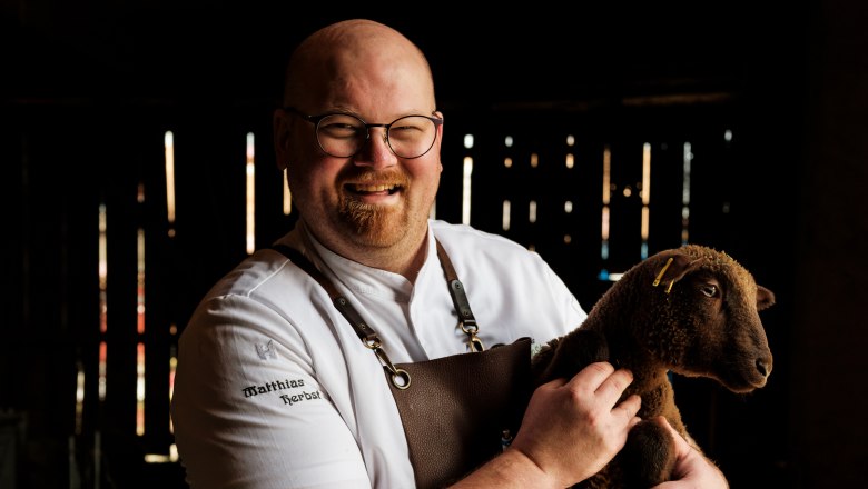 A man in chef's clothes holds a brown sheep in a stable.