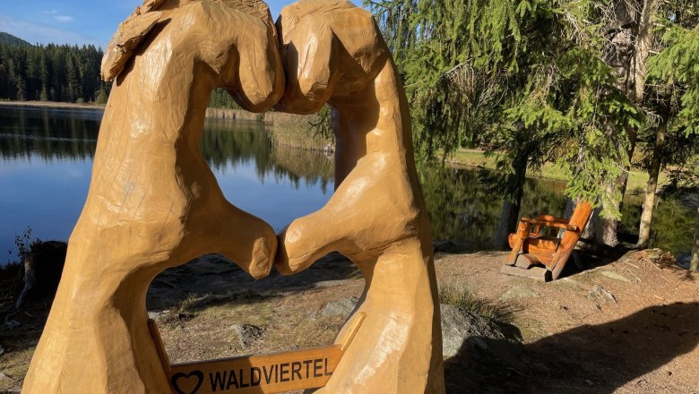 Wooden sculpture of two hands forming a heart with the inscription 'Waldviertel' in front of a lake and forest.