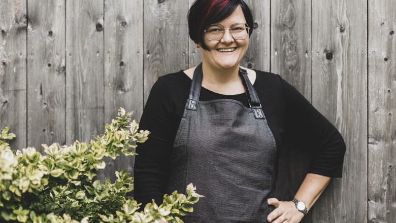 A woman with glasses and an apron stands smiling in front of a wooden wall next to a bush.