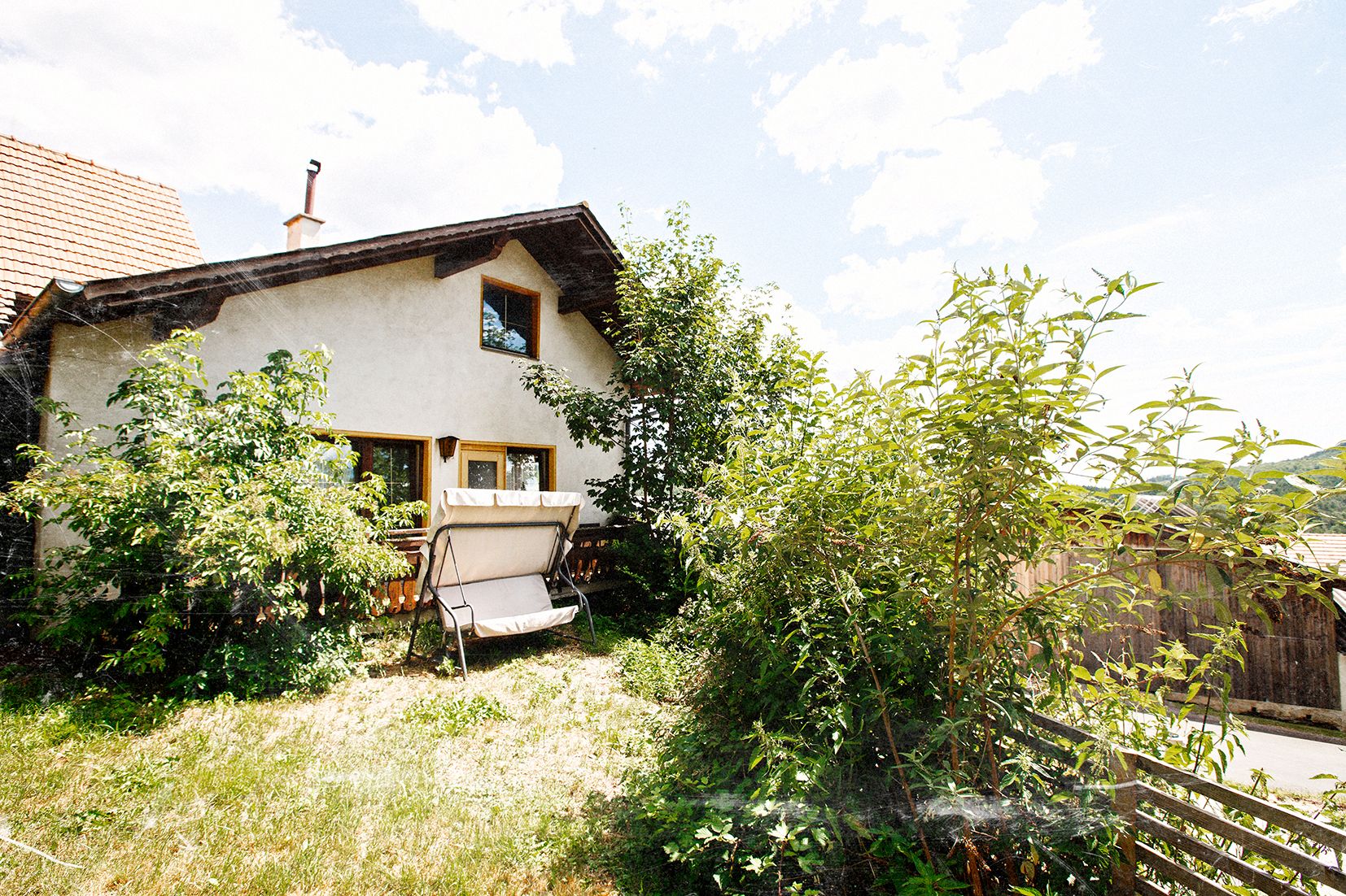A cozy house with a garden and a swing bench, surrounded by lush greenery and trees under a blue sky.