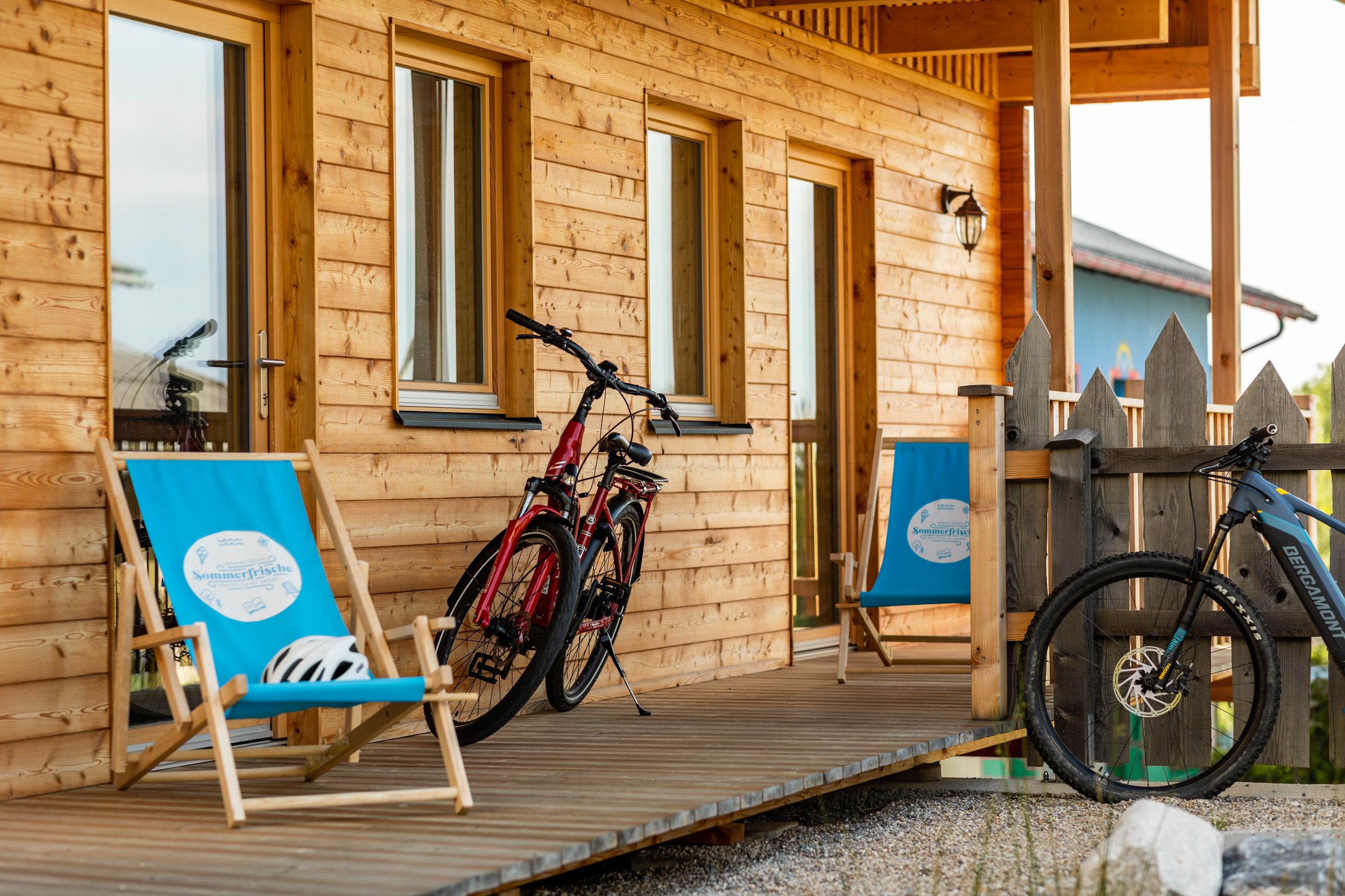 Wooden veranda with two bicycles and blue deckchairs in front of a wooden house.