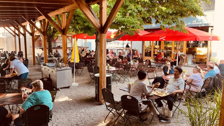 A beer garden with a wooden roof, red parasols and lots of people at tables.