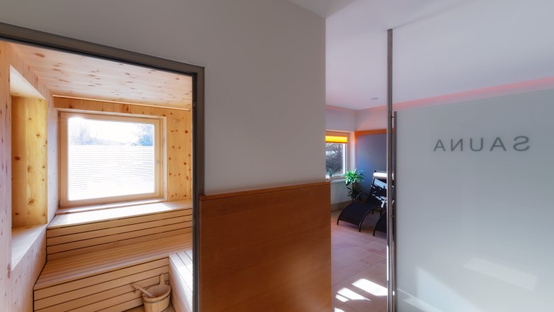 Interior view of a sauna with wooden paneling and window, next to it a relaxation room with loungers.