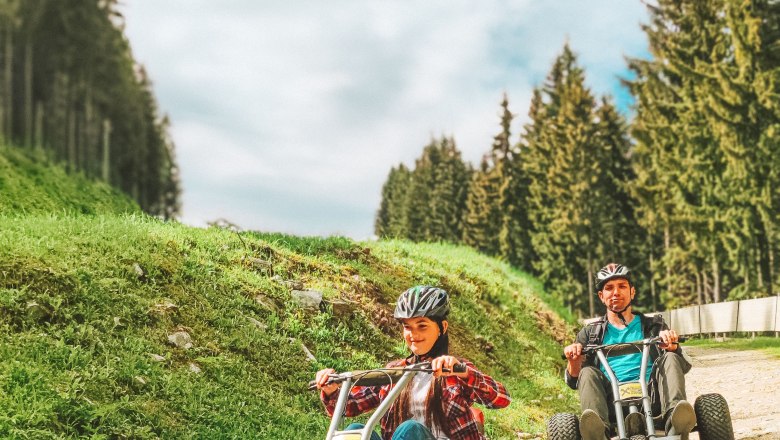 Two people ride mountain carts on a gravel road, surrounded by trees and blue sky.