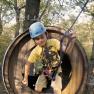 A child with helmet and climbing equipment in a wooden climbing park tunnel.