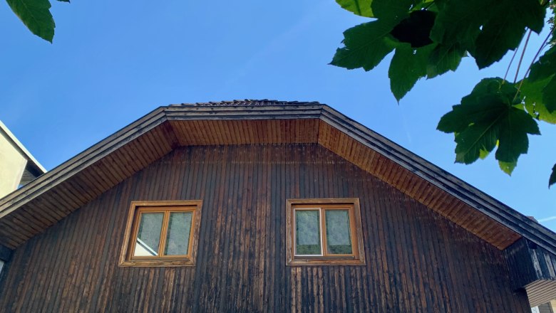 Attic of a house with wooden paneling and two windows, surrounded by green leaves and blue sky.