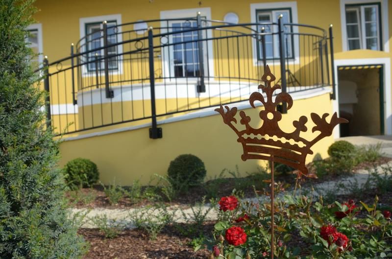 A yellow building with a balcony and a garden with red roses and a metal crown as decoration.