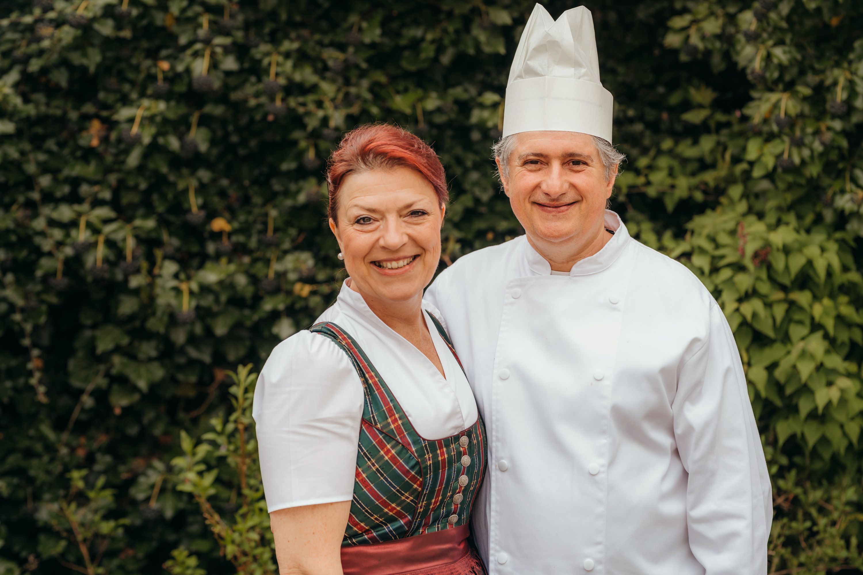 A man in chef's clothes and a woman in traditional costume smile at the camera against a backdrop of green leaves.