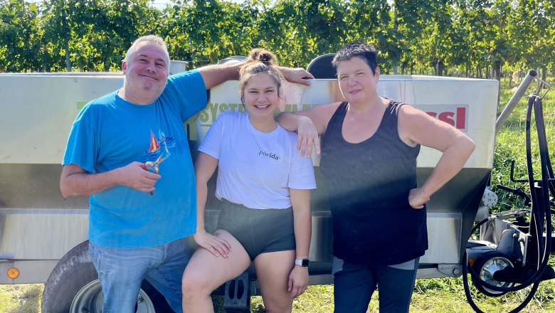 Three people are standing in front of a vineyard next to a trailer.