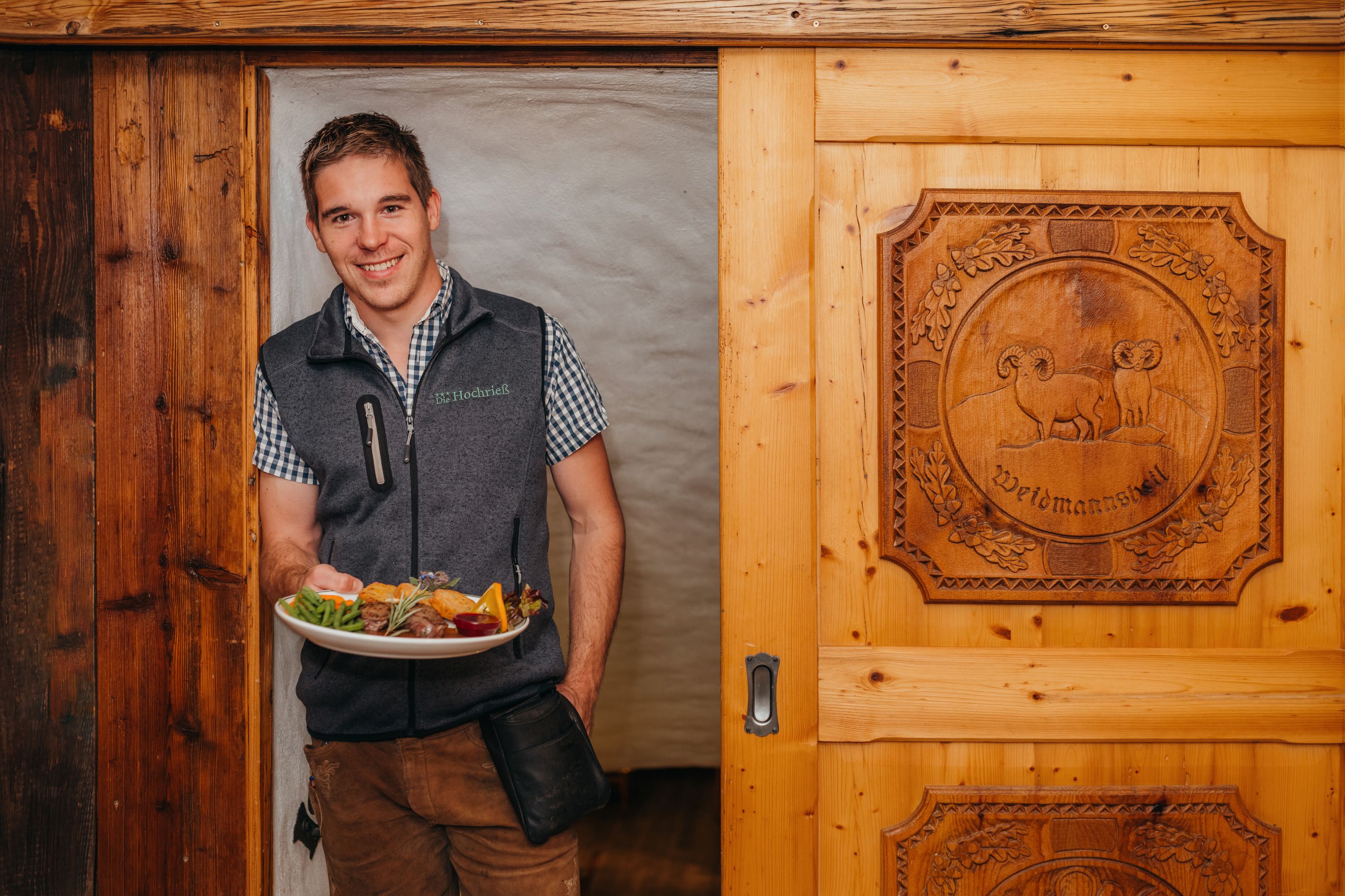A man in traditional dress holds a plate of food in front of a wooden door with a carved motif.