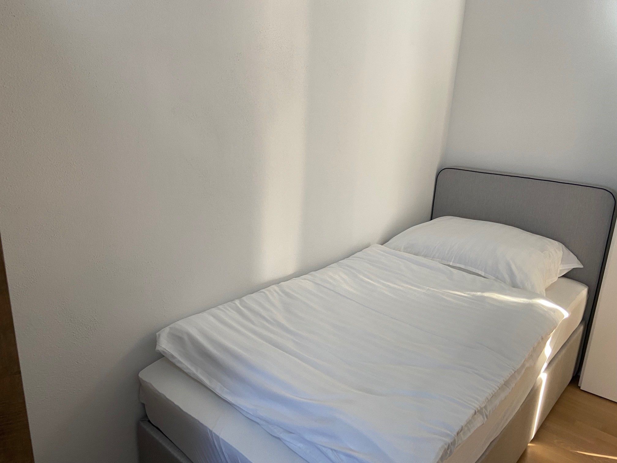 A simple, neatly made single bed in a bright room with white walls.