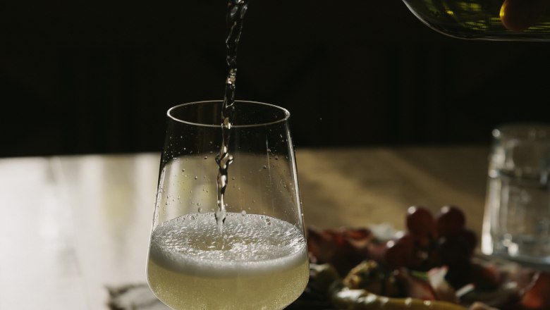 A glass of white wine is poured on a table, with a plate of snacks in the background.