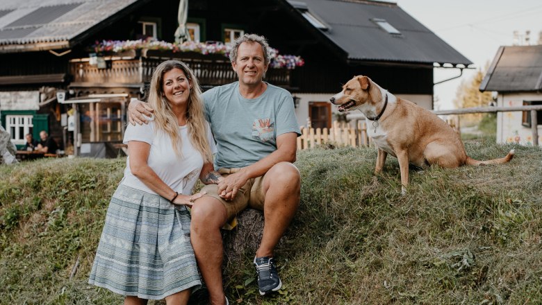A couple sits on a hill in front of a hut with a dog next to them.