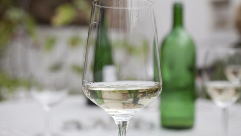 A wine glass with white wine in the foreground, blurred green wine bottle in the background.