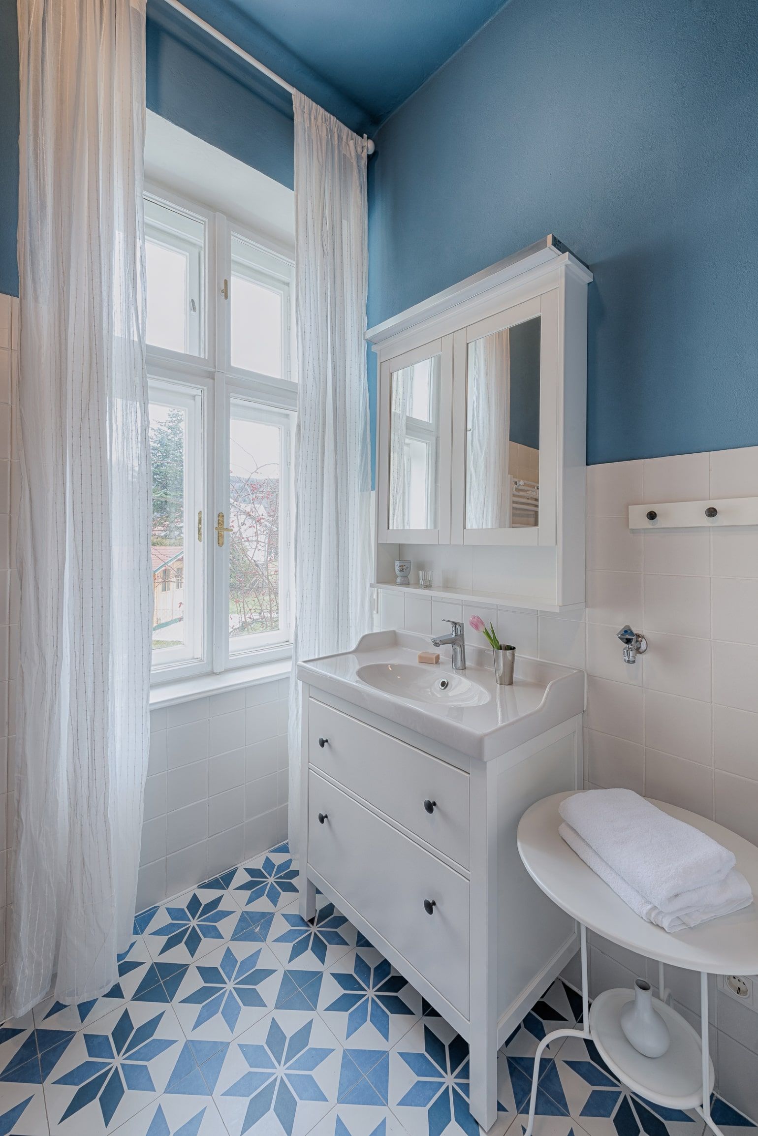 Bright bathroom with blue and white tiles, white washbasin and window with white curtains.