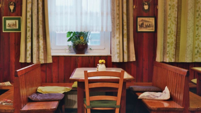 Cozy dining room with wooden furniture, table, chairs and bench, windows with curtains, plants and wall decorations.