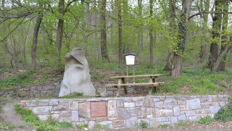A natural stone and a bench in the forest, surrounded by trees and a stone wall.